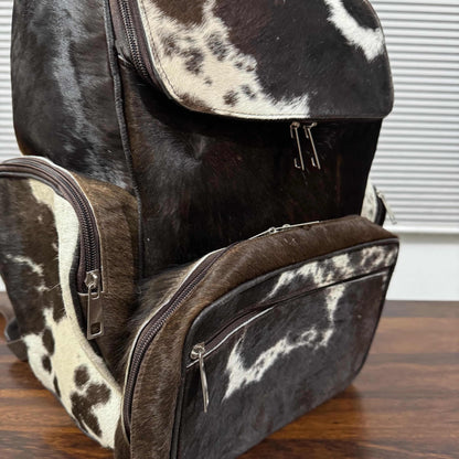 A structured backpack and matching diaper bag set made from dark brown and cream cowhide, displayed on a wooden tabletop with a window blind in the background.