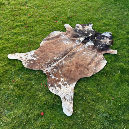 A natural cowhide rug with a mottled brown, black, and white pattern, laid flat on a green grass lawn.