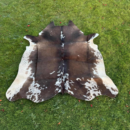 A natural cowhide rug with a brown and white spotted pattern, laid flat on a green grass lawn.