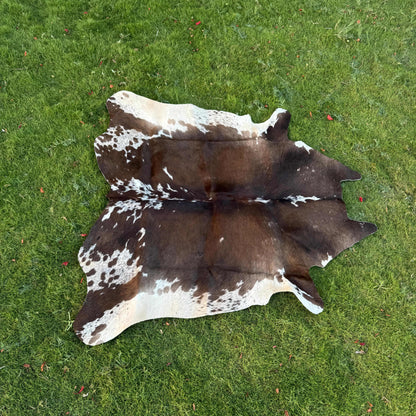 A natural cowhide rug with a brown and white spotted pattern laid flat on a green grass lawn.