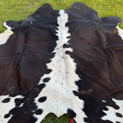A black and white cowhide rug laid flat on green grass, showing the natural hair-on-hide texture and spotted pattern.