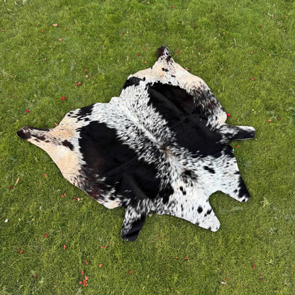 A natural cowhide rug with a black and white spotted pattern and tan patches, laid flat on a green grass lawn.