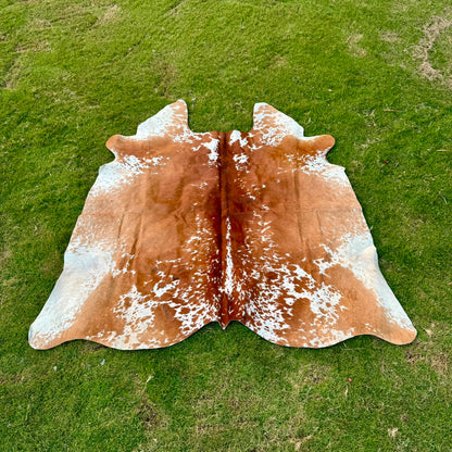 A natural cowhide rug with a brown and white speckled pattern, laid flat on a green grass lawn.