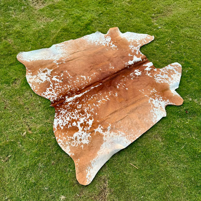 A natural cowhide rug with a brown and white spotted pattern, laid out on a green grass lawn.