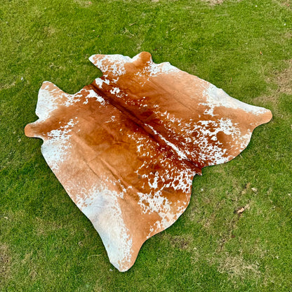 A natural cowhide rug with a brown and white mottled pattern laid out on a green grass lawn.