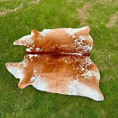 A natural cowhide rug with a brown and white spotted pattern laid out on a green grass lawn.