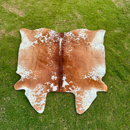 A natural cowhide rug with a brown and white mottled pattern, laid flat on a green grass lawn.