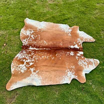 A natural cowhide rug with a brown and white spotted pattern, laid flat on a green grass lawn.