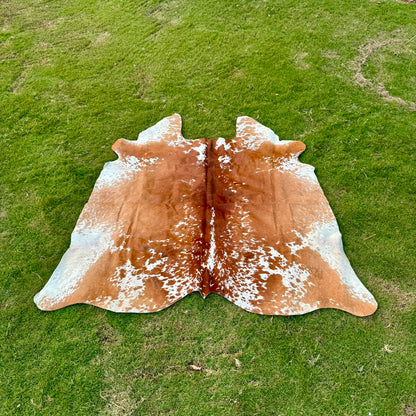 A natural cowhide rug with a brown and white mottled pattern laid out on a green grass lawn.