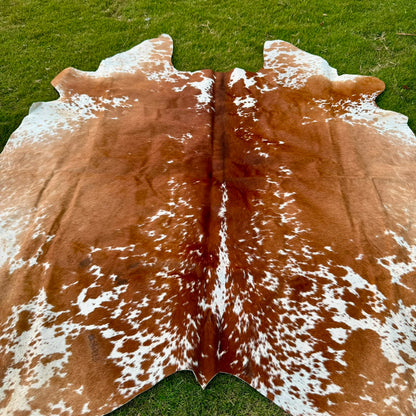 A natural cowhide rug with a brown and white spotted pattern laid out on green grass.