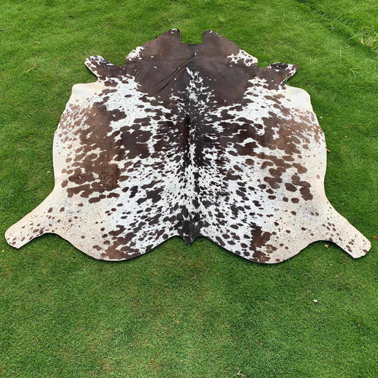 A natural cowhide rug with a brown and white spotted pattern, laid flat on a green grass lawn.
