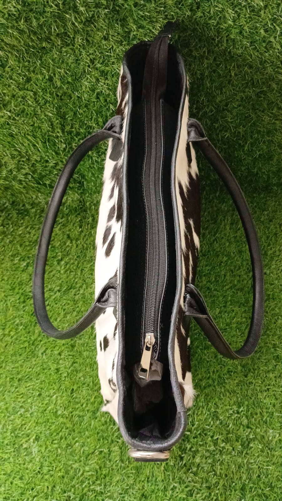 A cowhide shoulder bag with black leather handles and trim, shown from a top-down view on a green grass background.