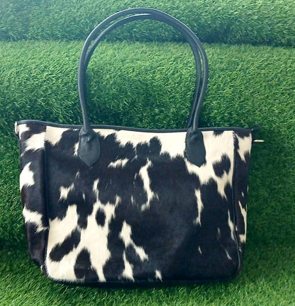 A black and white cow-print handbag with black leather handles, displayed on a green grass background.