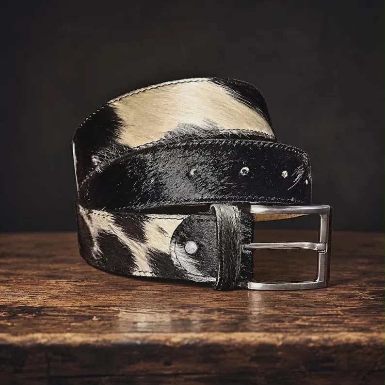 A black and cream cowhide belt with a silver-tone metal buckle, coiled on a rustic wooden surface against a dark background.