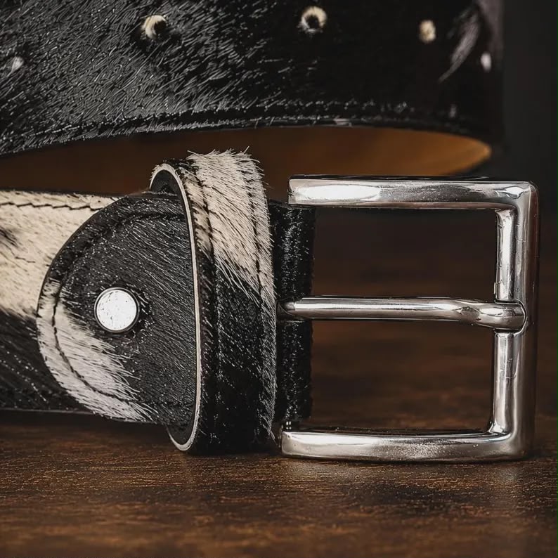 A close-up of a western-style belt with a black and white cowhide pattern, a polished silver buckle, and silver rivets, resting on a dark wooden surface.