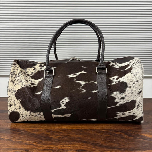 A duffle bag with a brown and white cowhide pattern and black faux-leather handles, resting on a wooden table in front of a gray slatted background.