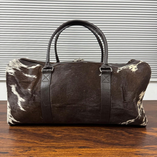 A dark brown and black cowhide duffel bag with black leather handles, resting on a wooden table in front of a gray slatted wall.