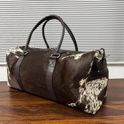 A duffle bag made of dark brown and black cowhide with black leather handles and straps, resting on a wooden table in front of a window with horizontal blinds.