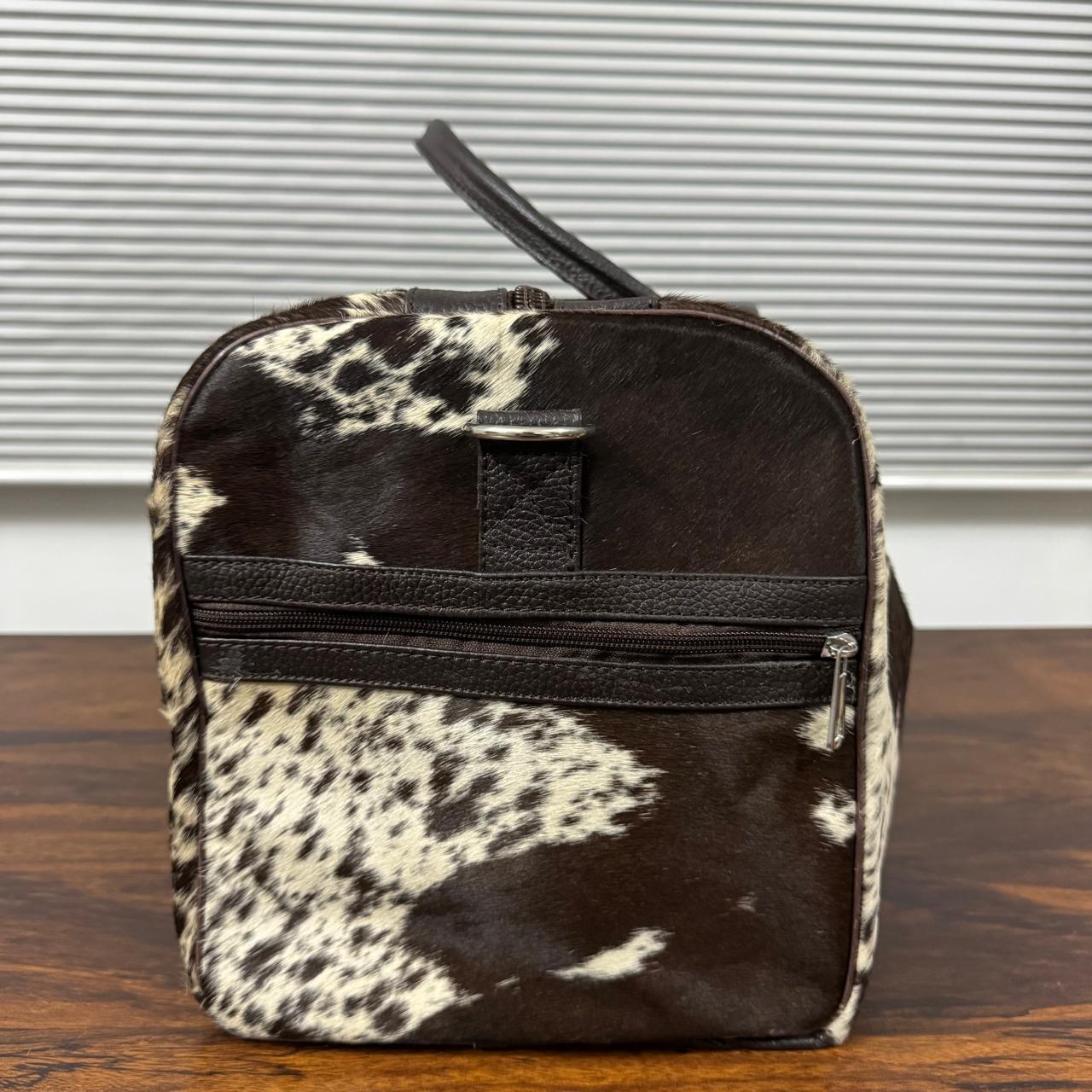 A small zippered toiletry bag with a black and white cowhide pattern, featuring a black leather handle and a horizontal black zipper, set on a wooden table in front of a window with blinds.