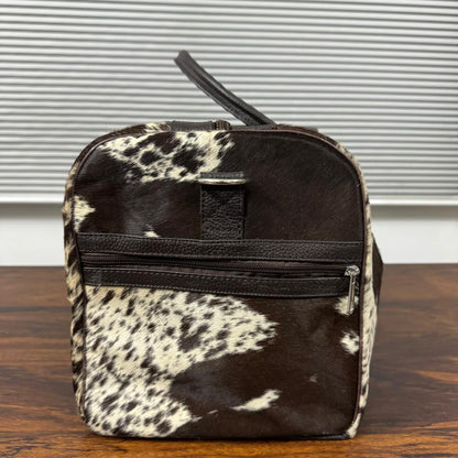 A small zippered toiletry bag with a black and white cowhide pattern, featuring a black leather handle and a horizontal black zipper, set on a wooden table in front of a window with blinds.