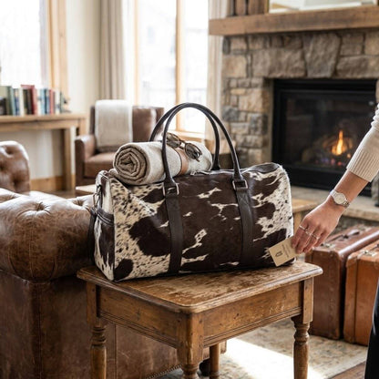 A cowhide-patterned duffel bag with dark brown leather handles sits on a wooden table in a cozy living room. A rolled blanket rests on top of the bag, and a person's hand is visible holding the bag's side tag.