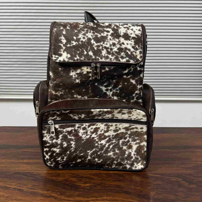 A cowhide-patterned backpack with dark brown leather trim, resting on a dark wooden table in front of a gray slatted window background.