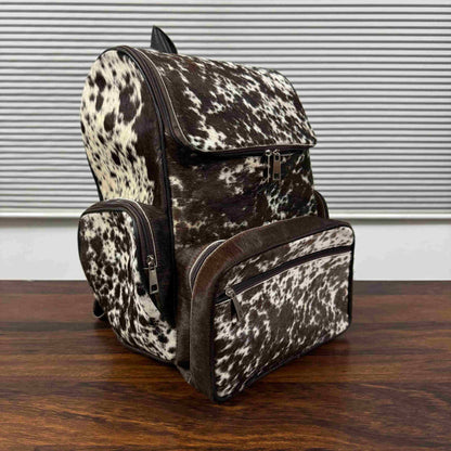 A cowhide-patterned backpack with matching front and side pouches, displayed on a wooden table in front of a window with horizontal blinds.