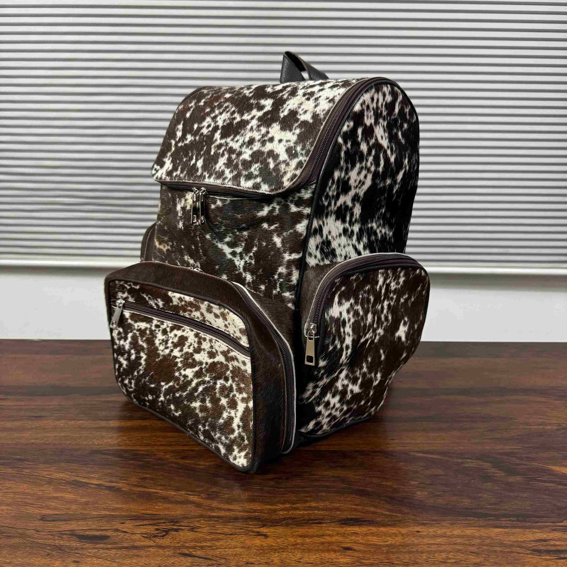 A cowhide-patterned backpack with matching zippered pouches, displayed on a wooden table in front of a window with horizontal blinds.