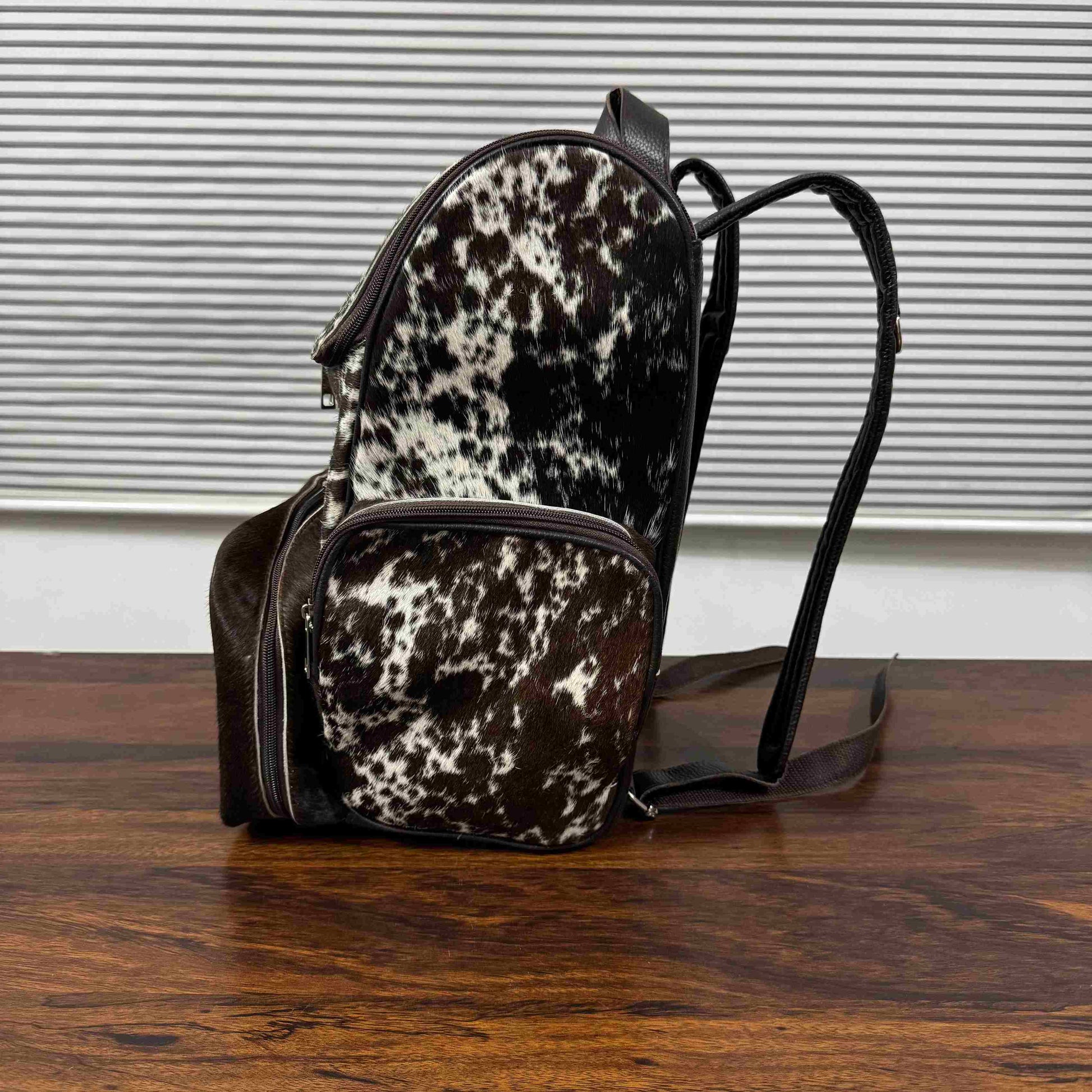 A small backpack with a black and white cowhide pattern and dark brown faux-leather trim, resting on a wooden table in front of a window with horizontal blinds.