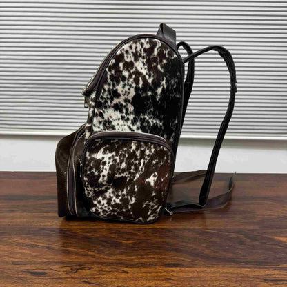 A small backpack with a black and white cowhide pattern and dark brown faux-leather trim, resting on a wooden table in front of a window with horizontal blinds.