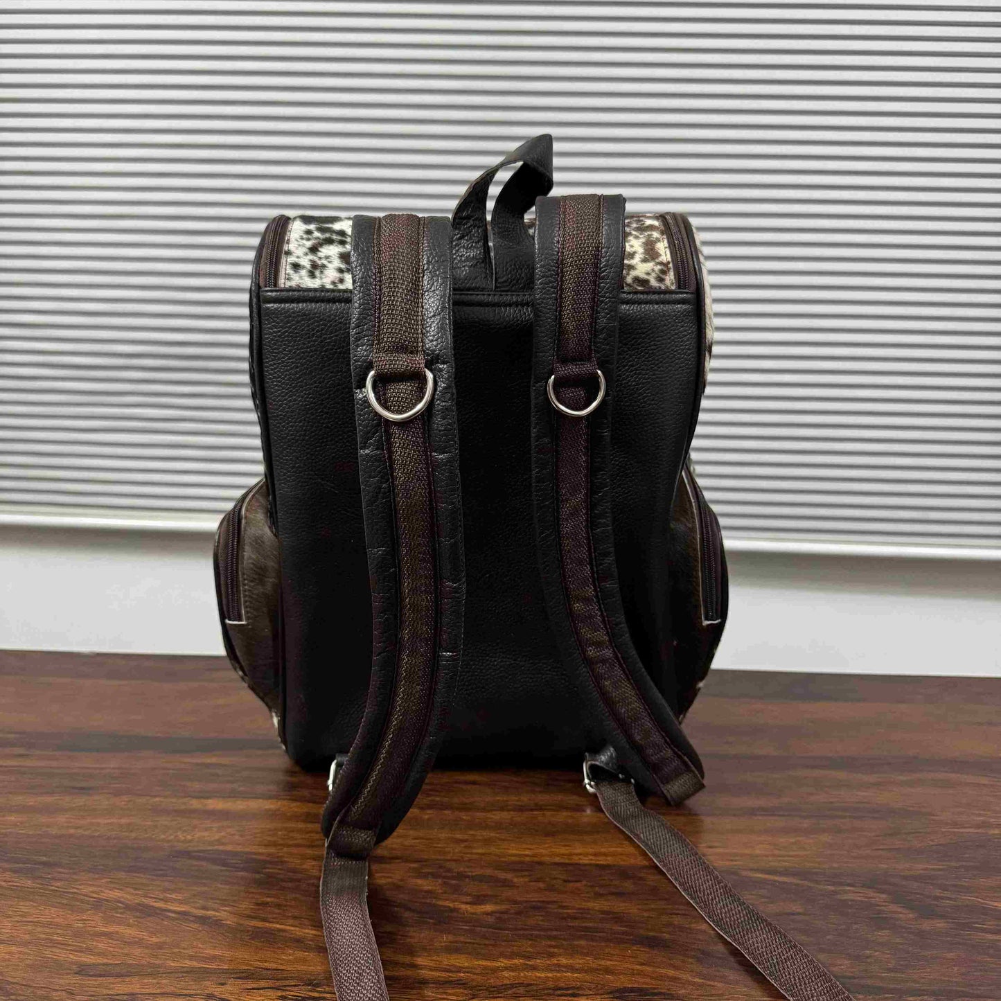 A black leather backpack with dark brown leather straps and leopard print accents on the top corners, shown from the back on a wooden surface in front of a window with horizontal blinds.