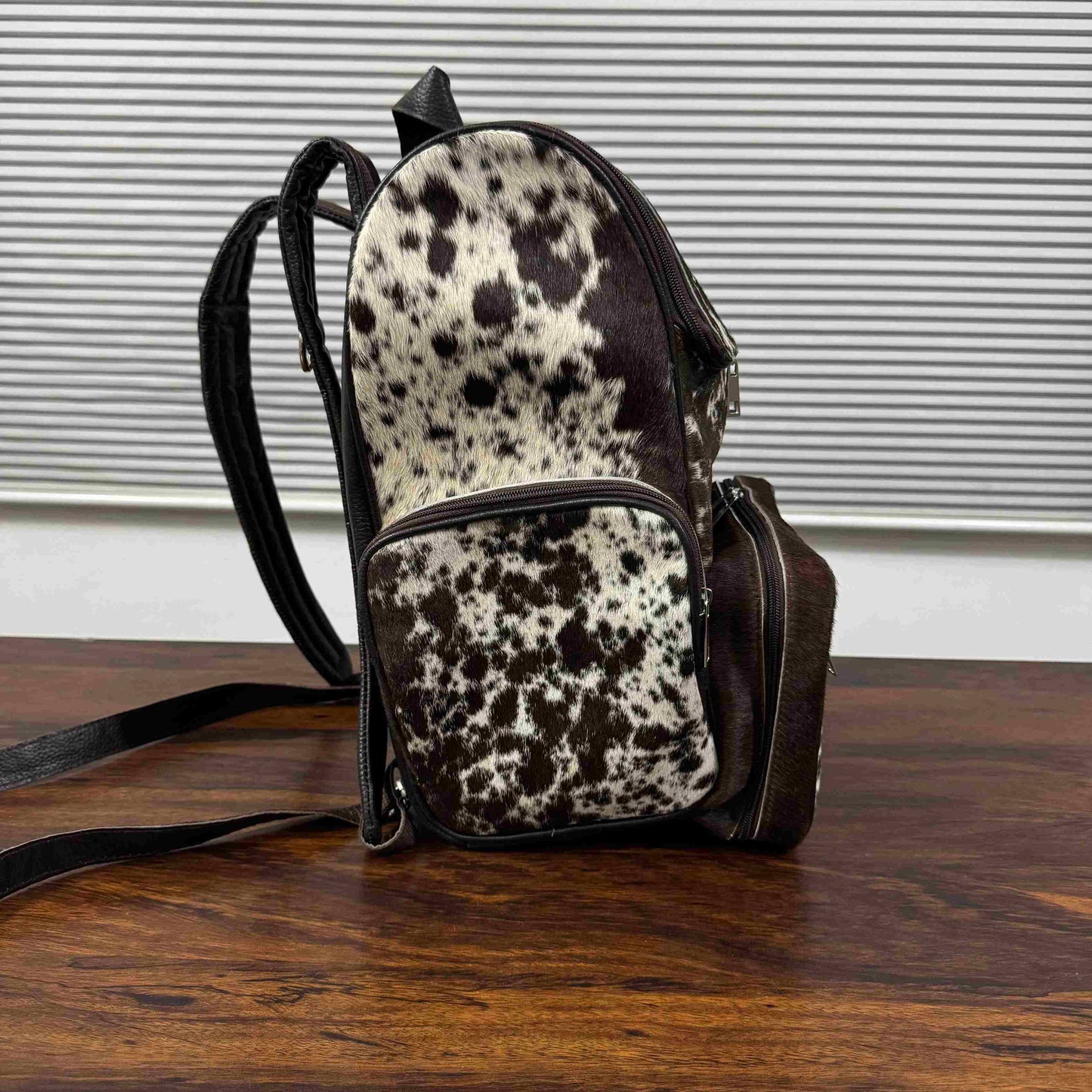 A small backpack with a cowhide pattern in brown and white, featuring dark brown faux-leather trim and straps, resting on a wooden table in front of a window with horizontal blinds.