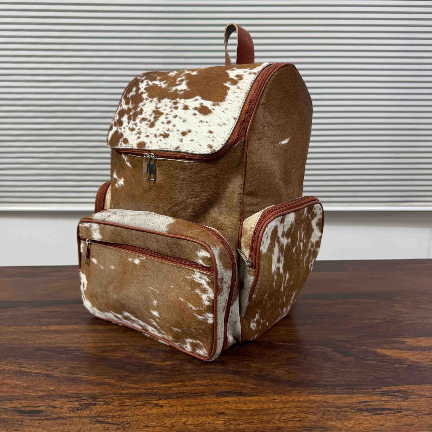 A brown and white cowhide-patterned backpack with reddish-brown trim, shown standing on a wooden table in front of a window with horizontal blinds.