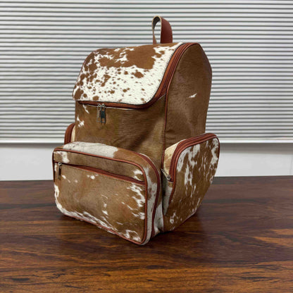 A brown and white cowhide-patterned backpack with reddish-brown trim, shown standing on a wooden table in front of a window with horizontal blinds.