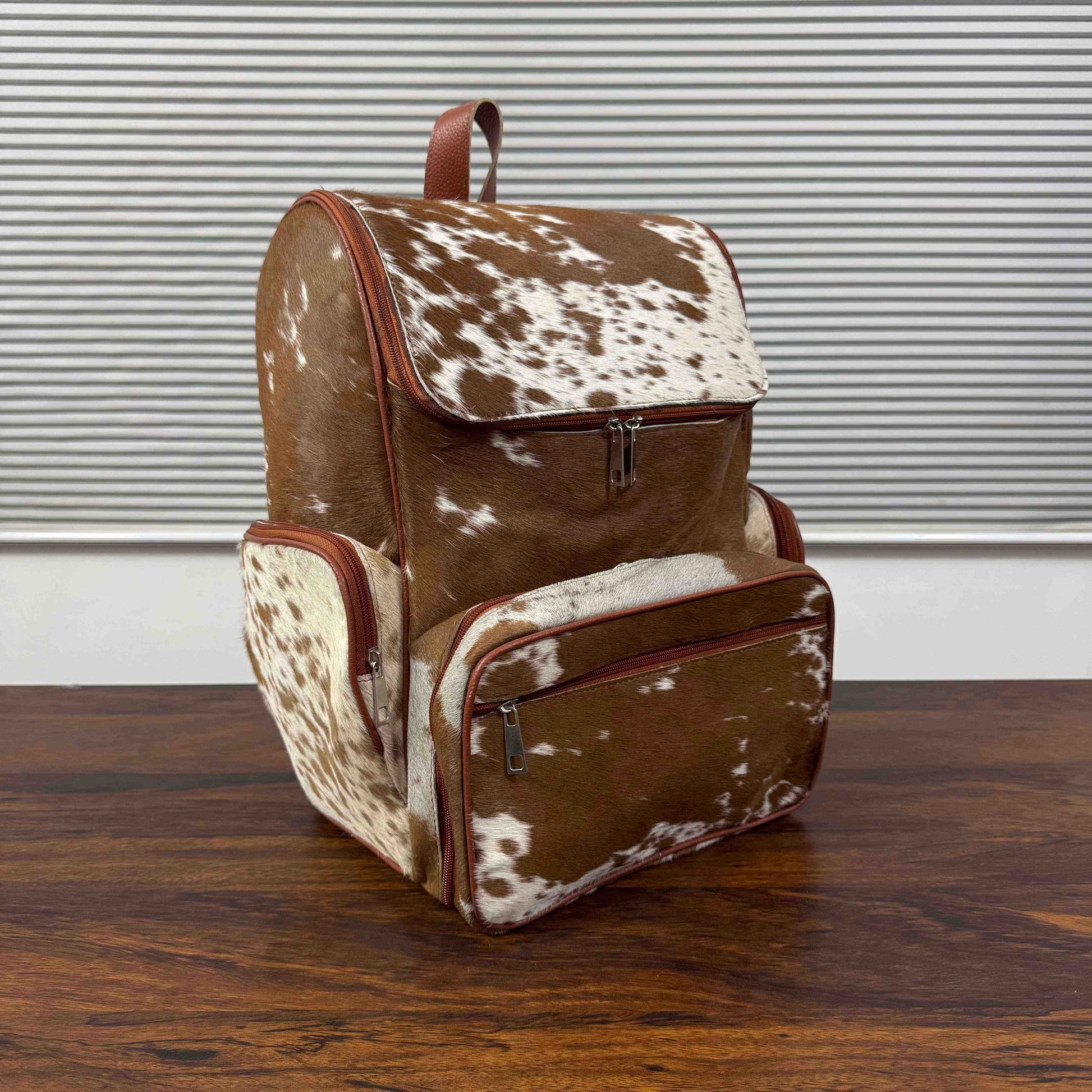 A brown and white cowhide-patterned backpack with brown leather trim, standing on a dark wooden tabletop in front of a gray slatted wall.