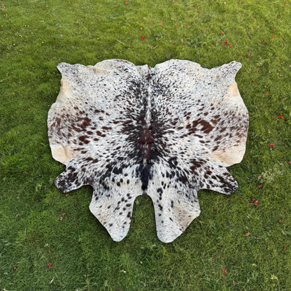 A natural cowhide rug with a black and white spotted pattern and tan edges, laid flat on a green grass lawn.