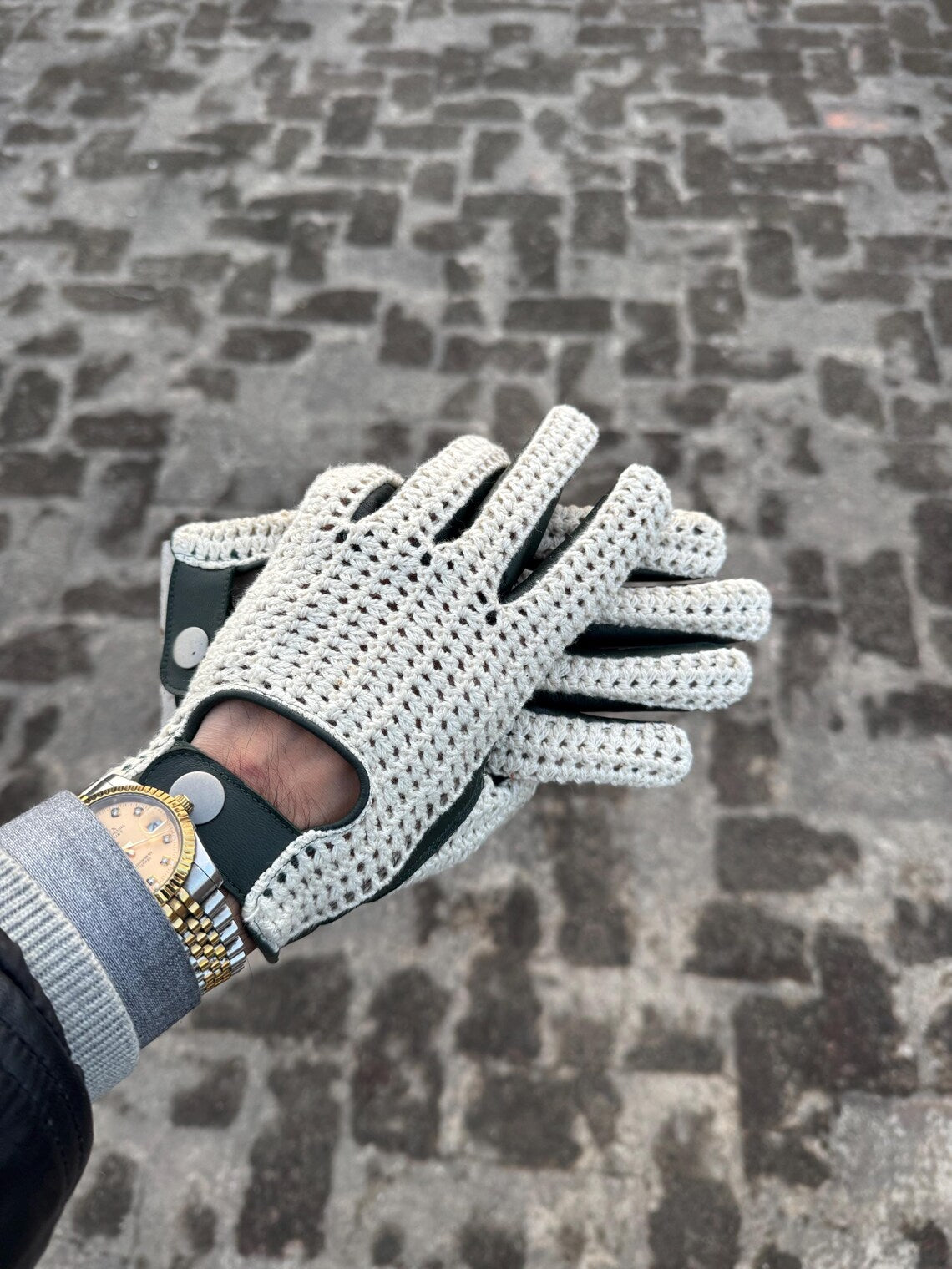 A close-up of a hand wearing a white knit golf glove with black accents and a gold watch, set against a blurred cobblestone pavement background.