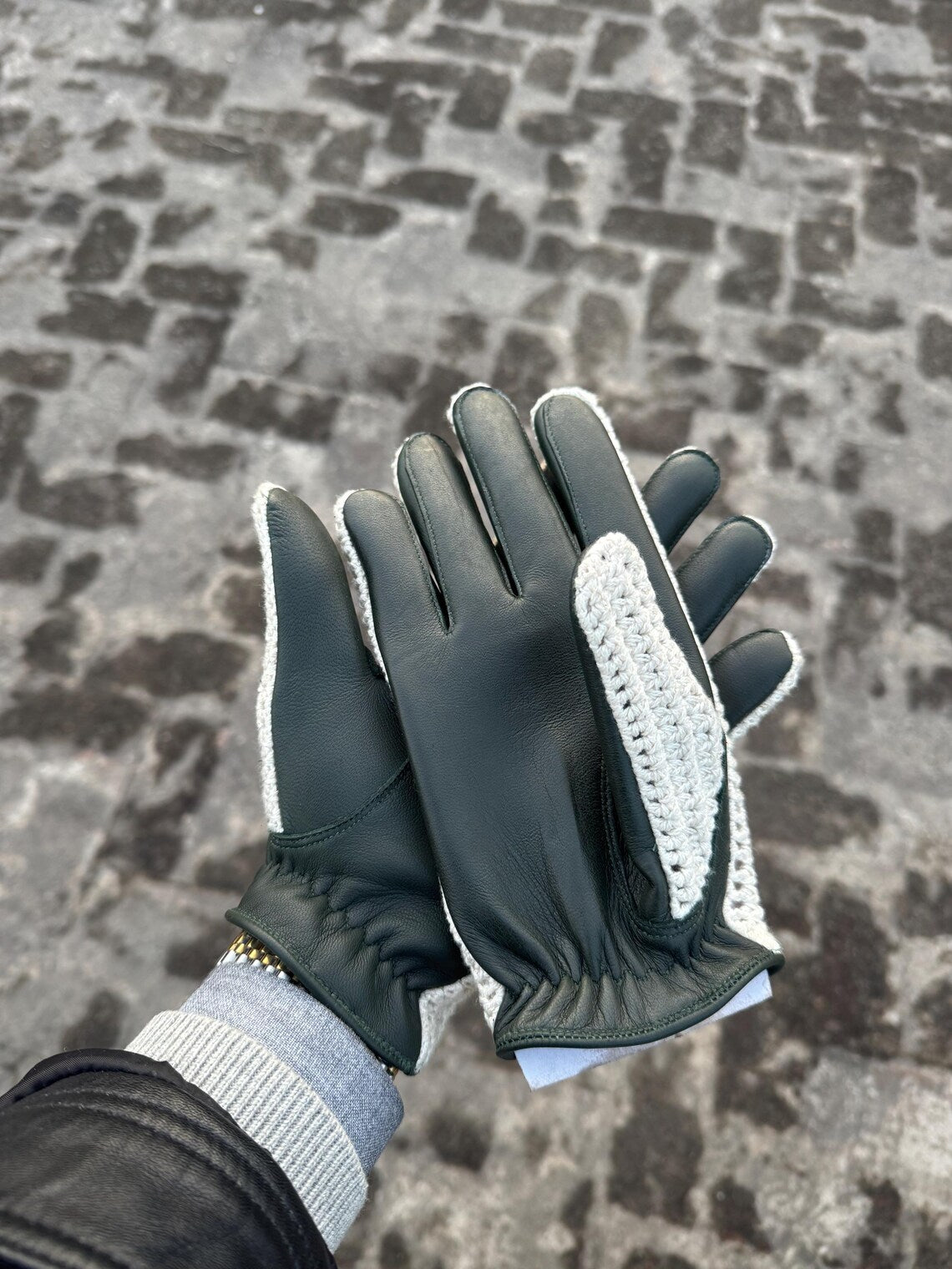 A pair of dark green leather gloves with white knit trim and textured white grip panels on the palms, shown being worn with a gray knit cuff and black jacket sleeve against a cobblestone pavement background.