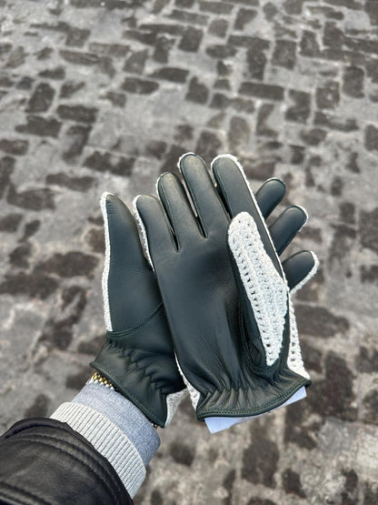 A pair of dark green leather gloves with white knit trim and textured white grip panels on the palms, shown being worn with a gray knit cuff and black jacket sleeve against a cobblestone pavement background.
