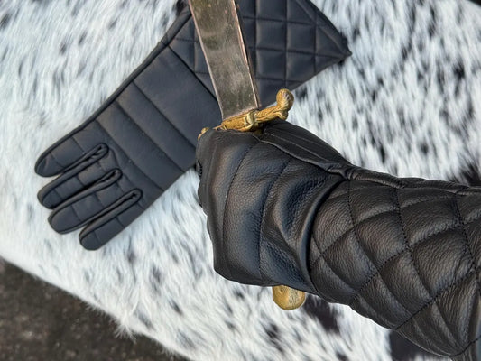 A close-up of a hand wearing a black quilted leather glove and holding a dagger with a gold hilt, with another black quilted glove laid out on a black-and-white spotted fur surface.