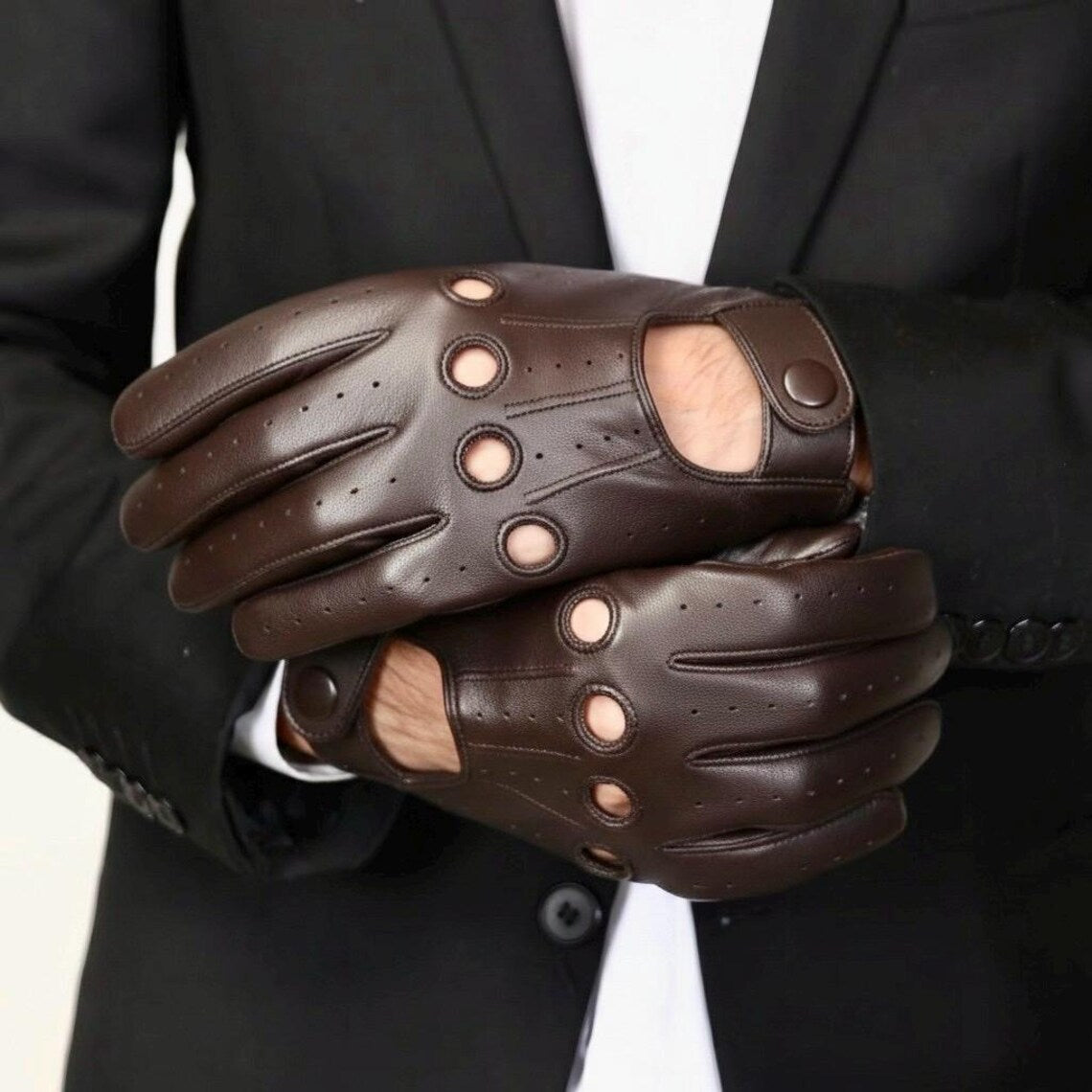 A pair of dark brown leather motorcycle gloves with circular cutouts on the back of the hand, worn on a model's hands in a black suit jacket.