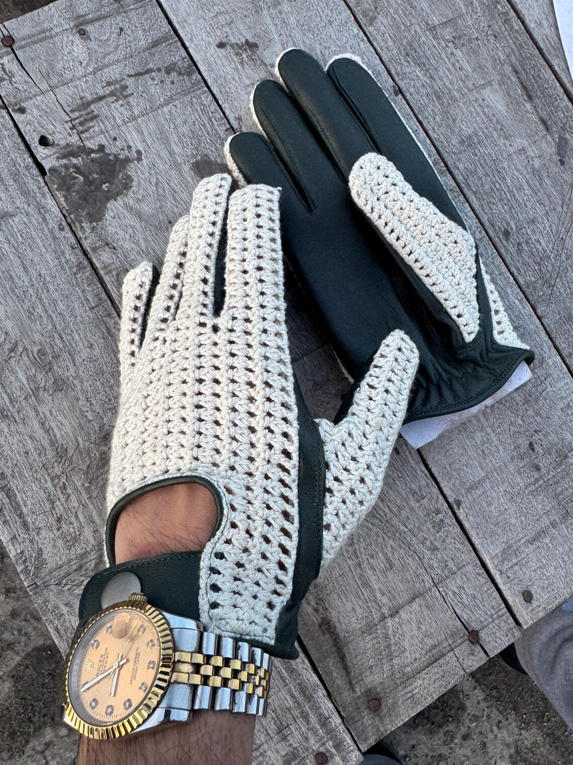 A pair of golf gloves, one cream crocheted with dark green trim and one solid dark green, shown on a person's hands resting on a weathered wooden surface.
