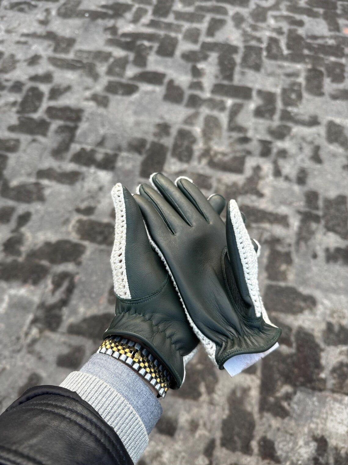 A pair of dark green leather gloves with white knit trim on the back of the hands, shown being worn with a gray knit cuff and a black leather jacket sleeve, set against a gray cobblestone pavement background.