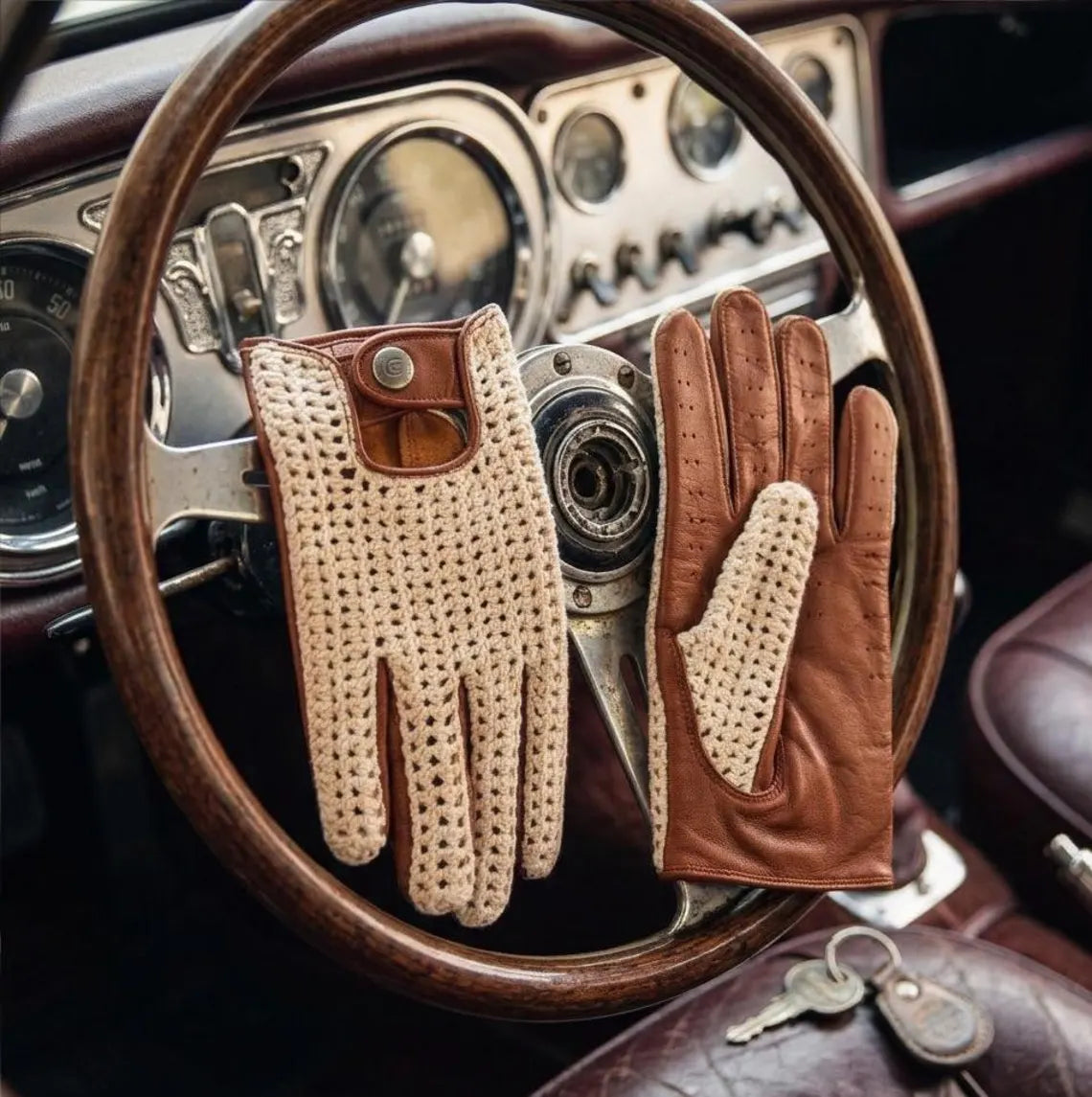 A pair of leather driving gloves with perforated cream panels and brown leather sections, displayed on a vintage wooden steering wheel inside a car.