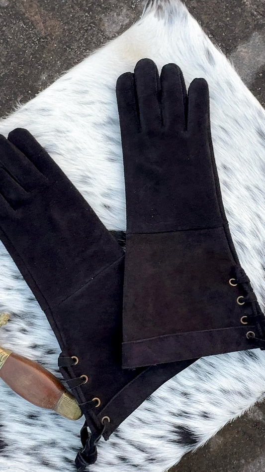 A pair of black suede opera gloves with brass eyelet lace-up cuffs, displayed on a white and black cowhide rug.