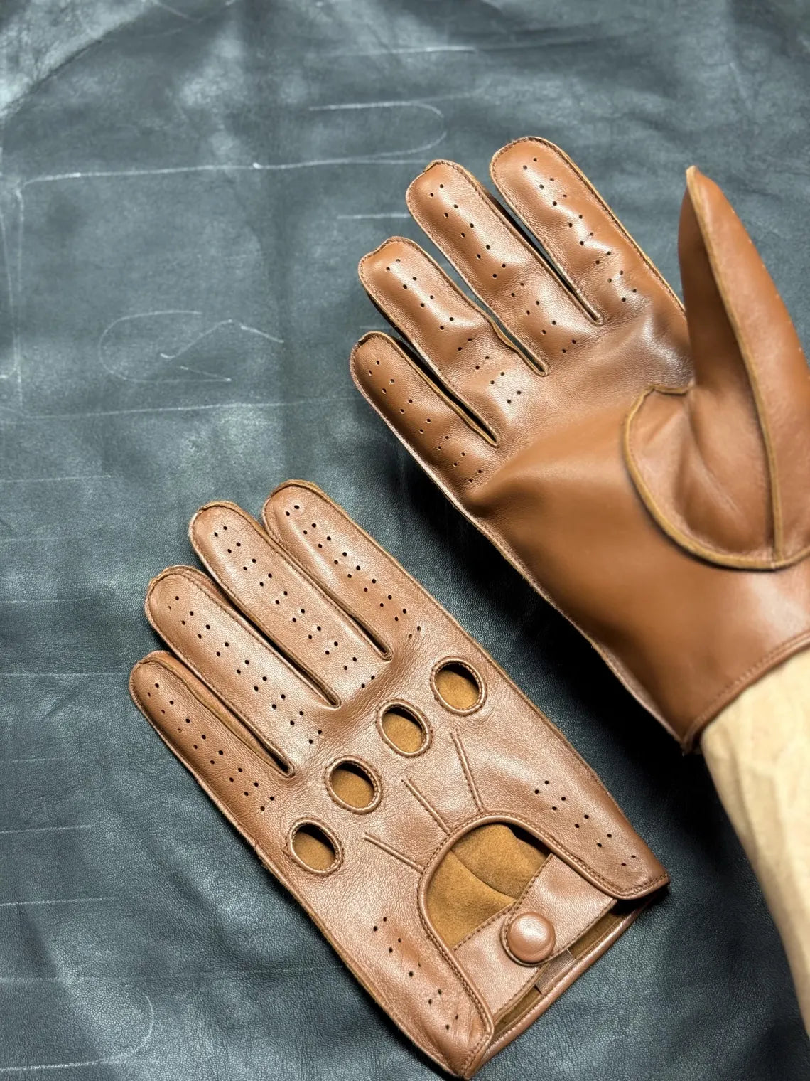 A pair of tan leather driving gloves, one shown palm-side and the other back-side, resting on a dark textured surface. The gloves feature perforated details on the fingers and palm, and a button closure on the back cuff.