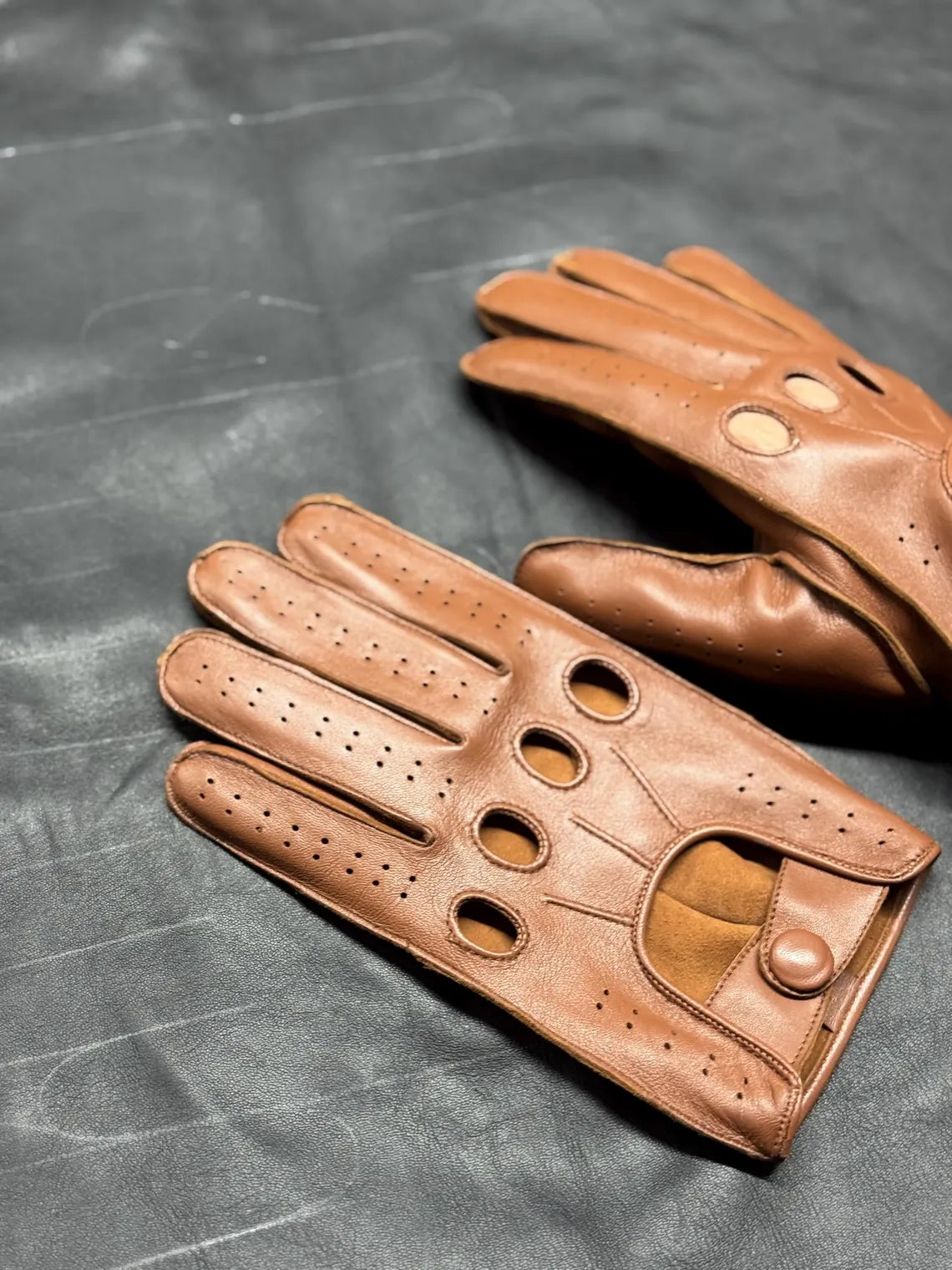A pair of brown leather driving gloves with perforated details and circular cutouts, displayed on a dark textured surface.