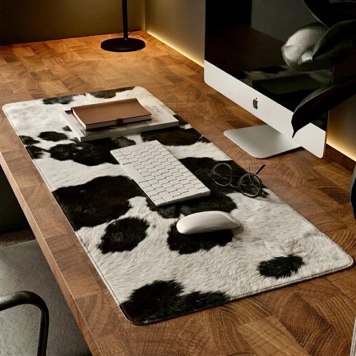 A black and white cow-print desk mat on a wooden desk, styled with a keyboard, mouse, glasses, and a notebook next to an iMac.