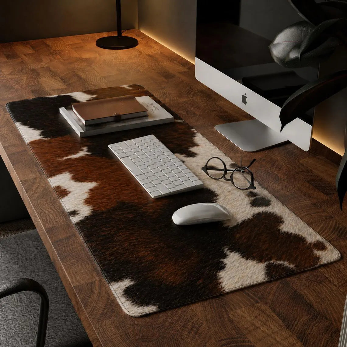 A rectangular desk mat with a brown and cream cowhide pattern is styled on a wooden desk, surrounded by a keyboard, mouse, notebook, glasses, and an iMac.