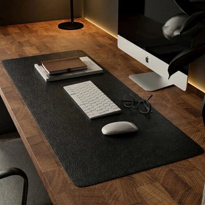 A large black desk mat with a textured surface is laid out on a wooden desk, styled with a white keyboard, white mouse, eyeglasses, and a notebook. An Apple iMac is partially visible in the background.